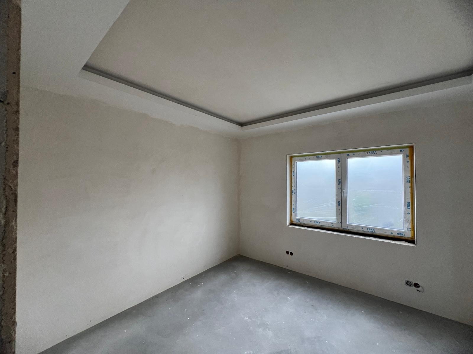 Pokój po szpachlowaniu ścian i wylewce podłogi. Widoczne nowe okno z folią ochronną i dekoracyjny sufit podwieszany z listwą. Stan surowy.