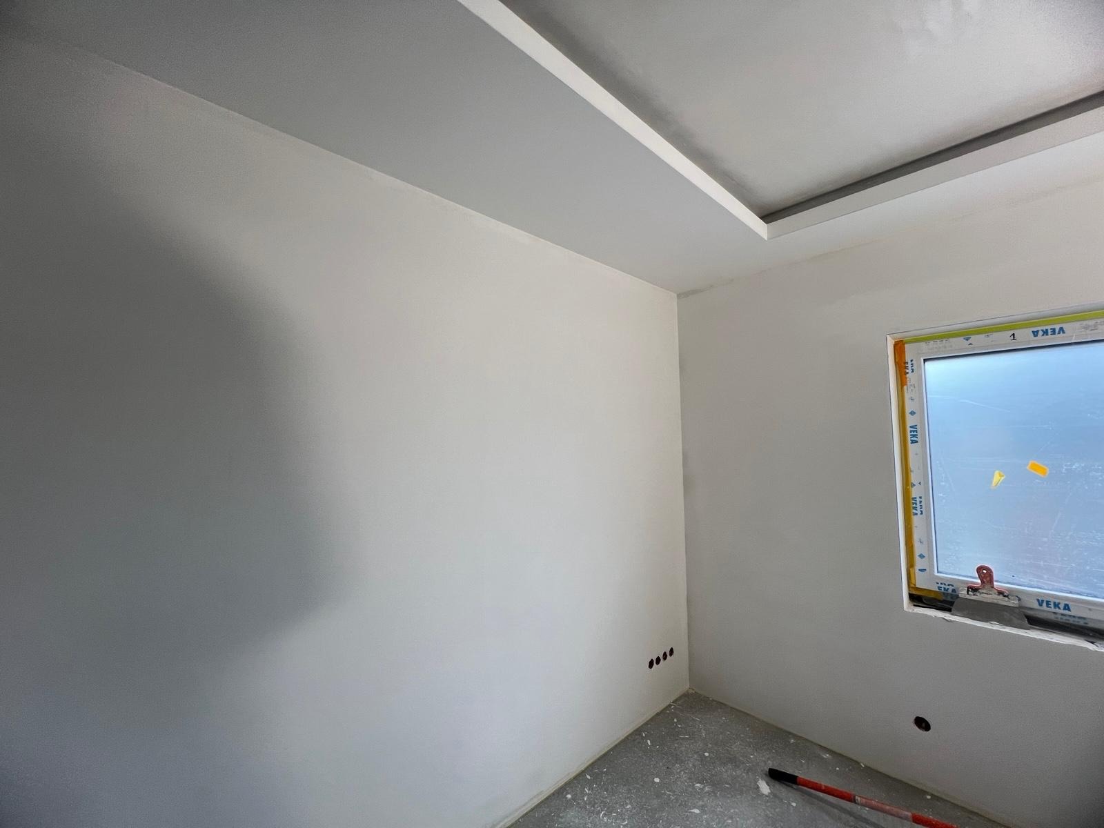 Gładkie, białe ściany po malowaniu w Koszalinie. Widoczne gniazdka elektryczne i okno z ramą. Na podłodze ślady farby i wałek malarski.