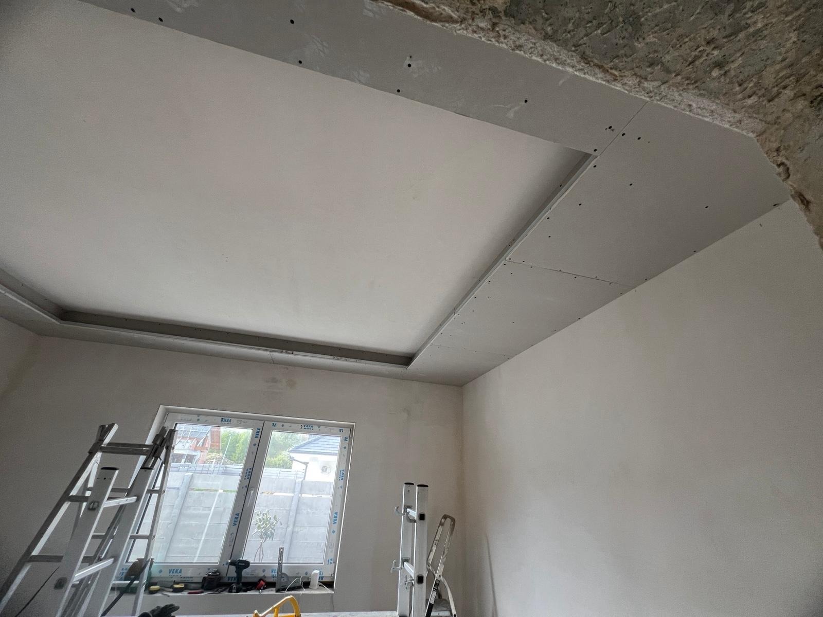 Montaż sufitu podwieszanego z płyt gipsowo-kartonowych w pomieszczeniu z oknem. Widoczne rusztowania i narzędzia. Prace wykończeniowe wnętrz w trakcie realizacji.
