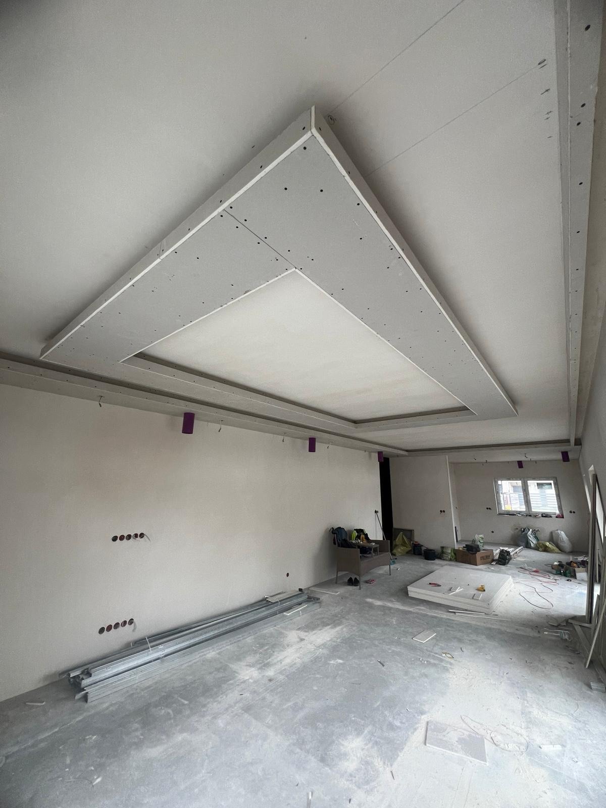 Sufit podwieszany z płyt karton-gips w kształcie trapezu, widoczne ślady po wkrętach. W tle surowe ściany i materiały budowlane na podłodze.