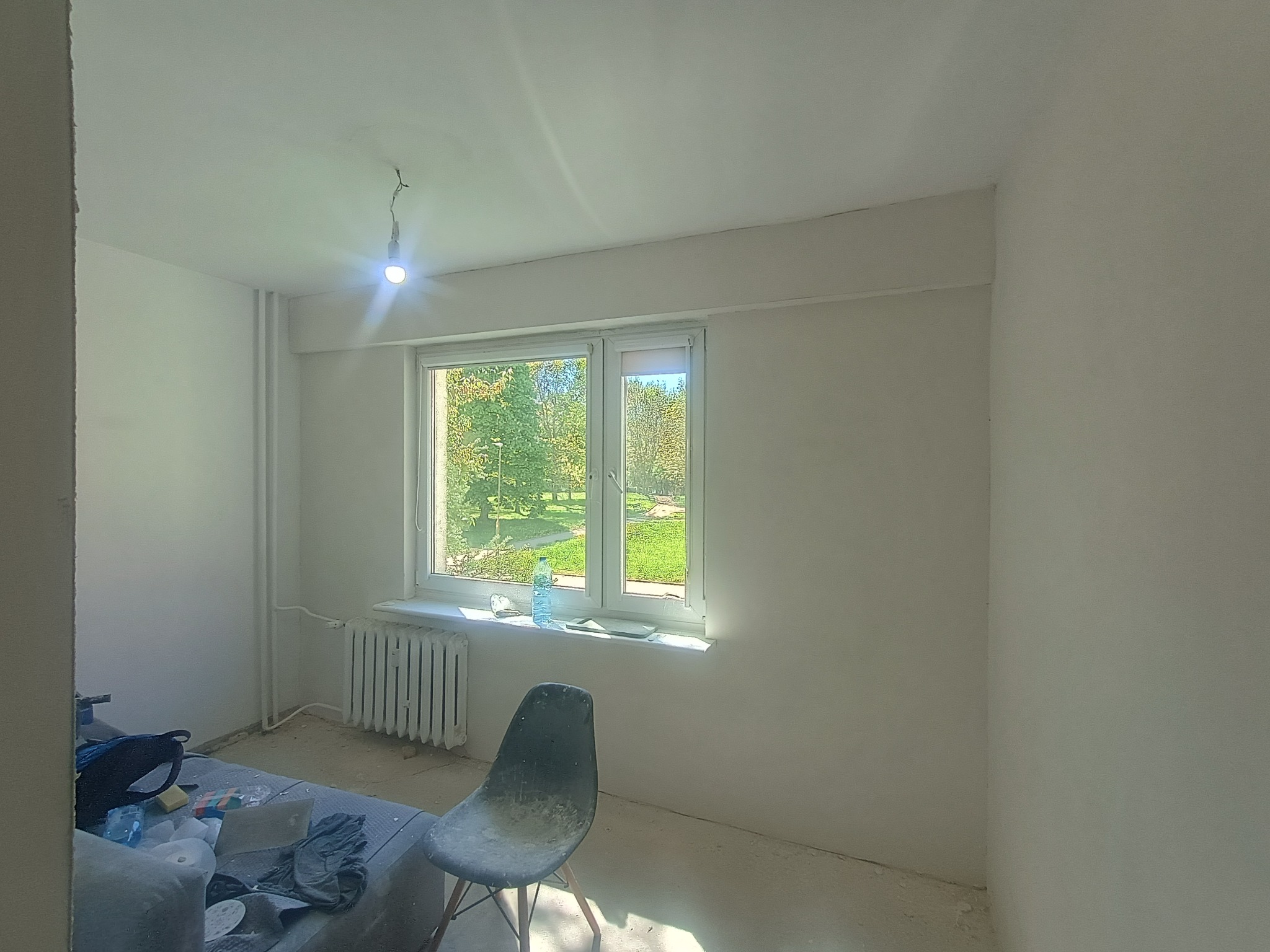 Wnętrze pokoju po szpachlowaniu ścian, przygotowane do malowania. Widoczne okno z widokiem na zieleń, grzejnik i krzesło. Ściany gładkie, białe, surowe oświetlenie.