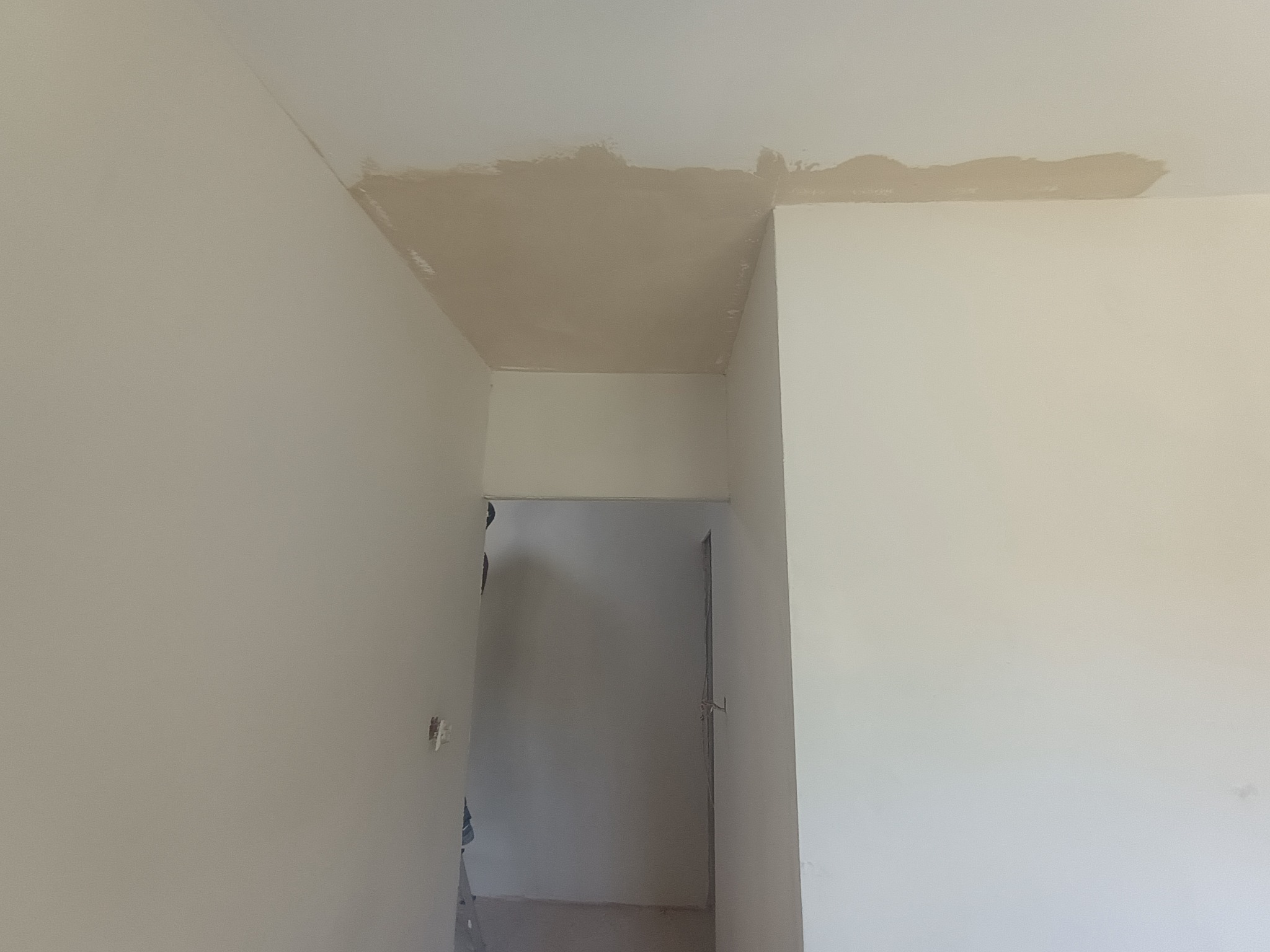 Wnętrze z widocznymi śladami prac wykończeniowych na suficie i ścianach, świeżo nałożona szpachla w rogu sufitu, przygotowanie do malowania, widok na sąsiednie pomieszczenie.