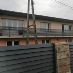 Nowoczesna balustrada balkonowa z poziomych profili, widoczna zza metalowego ogrodzenia panelowego w kolorze grafitowym, z betonowym słupkiem.