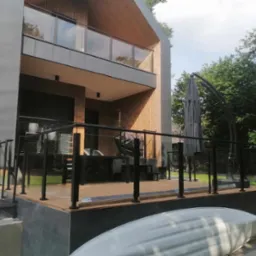Nowoczesny dom z drewnianą elewacją i balkonem zabezpieczonym szklaną balustradą z czarnymi słupkami, widok na taras z czarną balustradą i meblami ogrodowymi, częściowo zasłonięty przez rozłożony...