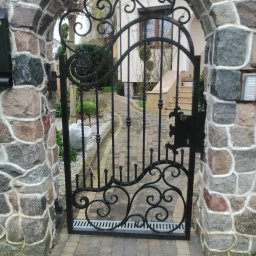 Łukasz Puchalski - Otwarta, ozdobna, czarna brama z kutego żelaza, osadzona w kamiennym łuku wjazdowym, prowadząca do ogrodu i domu.