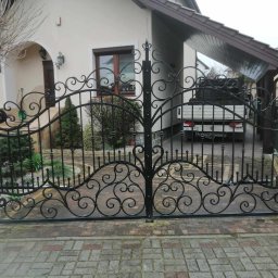 Łukasz Puchalski - Ozdobna, dwuskrzydłowa brama z kutego żelaza, z motywami roślinnymi, prowadząca do posesji z beżowym domem i wiatą garażową, widziana z poziomu brukowanego podjazdu.