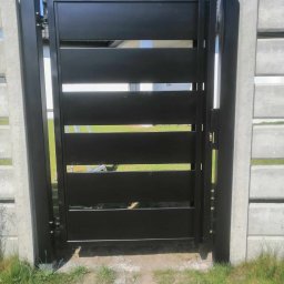 Łukasz Puchalski - Nowoczesna, czarna brama wjazdowa z poziomymi panelami, zamontowana pomiędzy betonowymi słupkami ogrodzeniowymi, widoczna z bliska w słoneczny dzień.