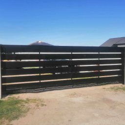 Łukasz Puchalski - Nowoczesna, przesuwna brama z poziomych, czarnych profili, osadzona na betonowych słupkach, na tle błękitnego nieba i zabudowy jednorodzinnej.