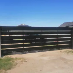 Nowoczesna, przesuwna brama z poziomych, czarnych profili, osadzona na betonowych słupkach, na tle błękitnego nieba i zabudowy jednorodzinnej.