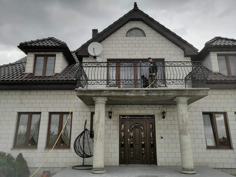 Mężczyzna myje balkon z kutą balustradą w dwupiętrowym domu z białej cegły, widoczny zwisający wąż i wiszące krzesło.