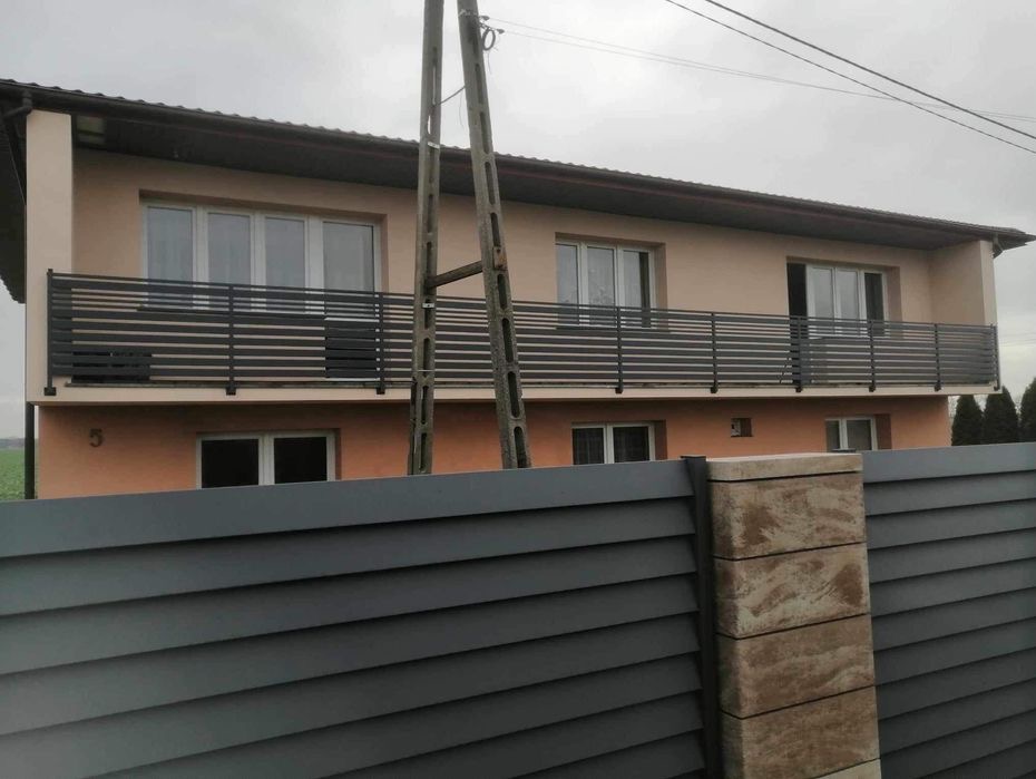 Nowoczesna balustrada balkonowa z poziomych profili, widoczna zza metalowego ogrodzenia panelowego w kolorze grafitowym, z betonowym słupkiem.