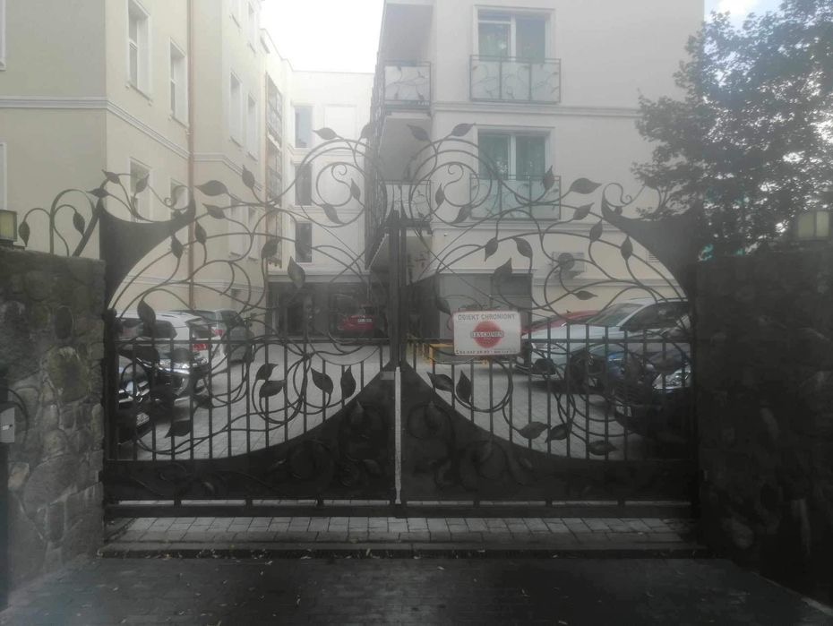 Dwuskrzydłowa brama wjazdowa z kutego metalu z motywem liści, zamontowana przy wejściu na posesję z budynkami mieszkalnymi; widoczny znak 'Obiekt Chroniony'.