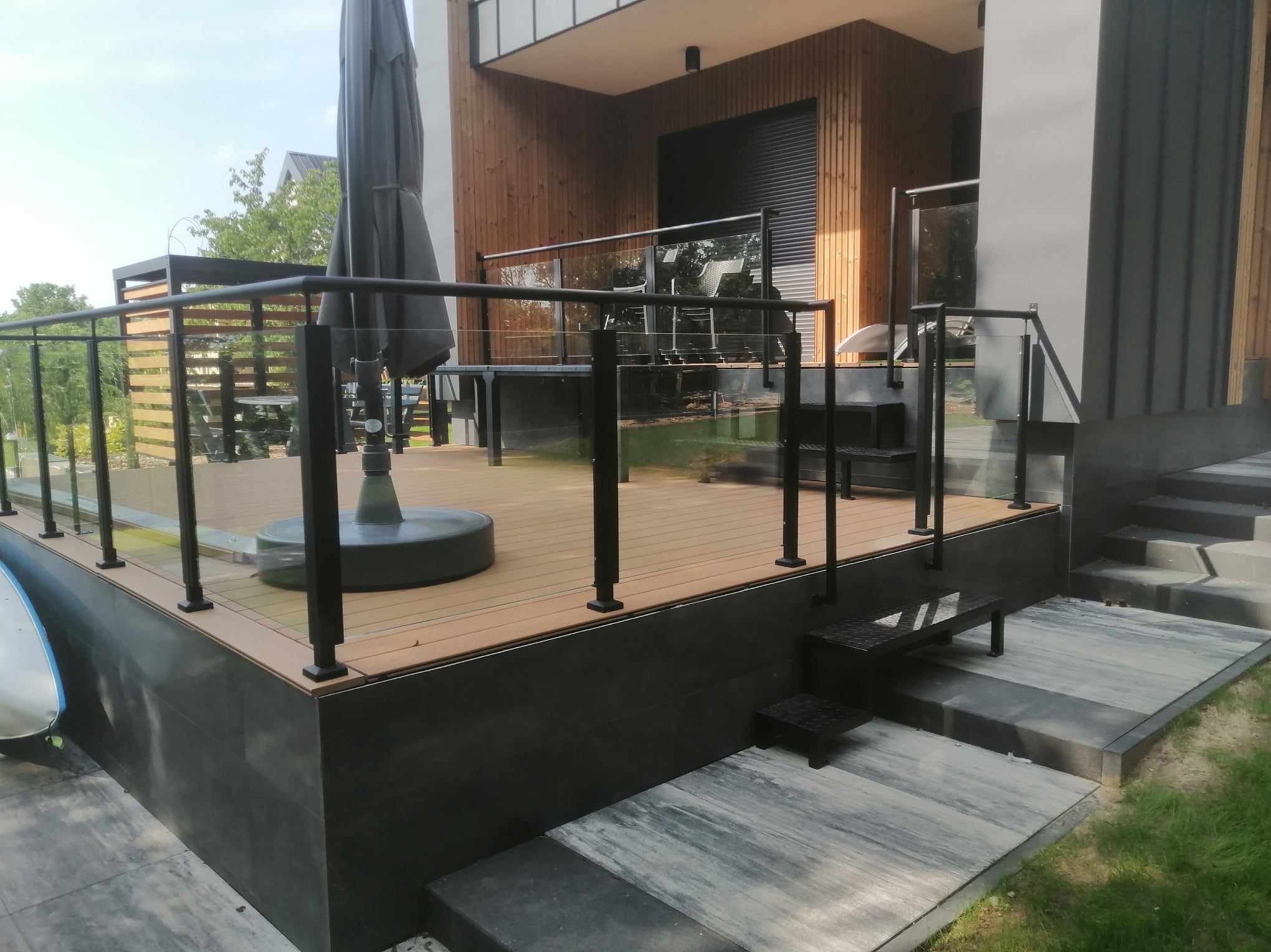 Nowoczesny taras z drewnianą podłogą i balustradami szklanymi z czarnymi słupkami, widok na dom z drewnianą elewacją i zewnętrznymi roletami.