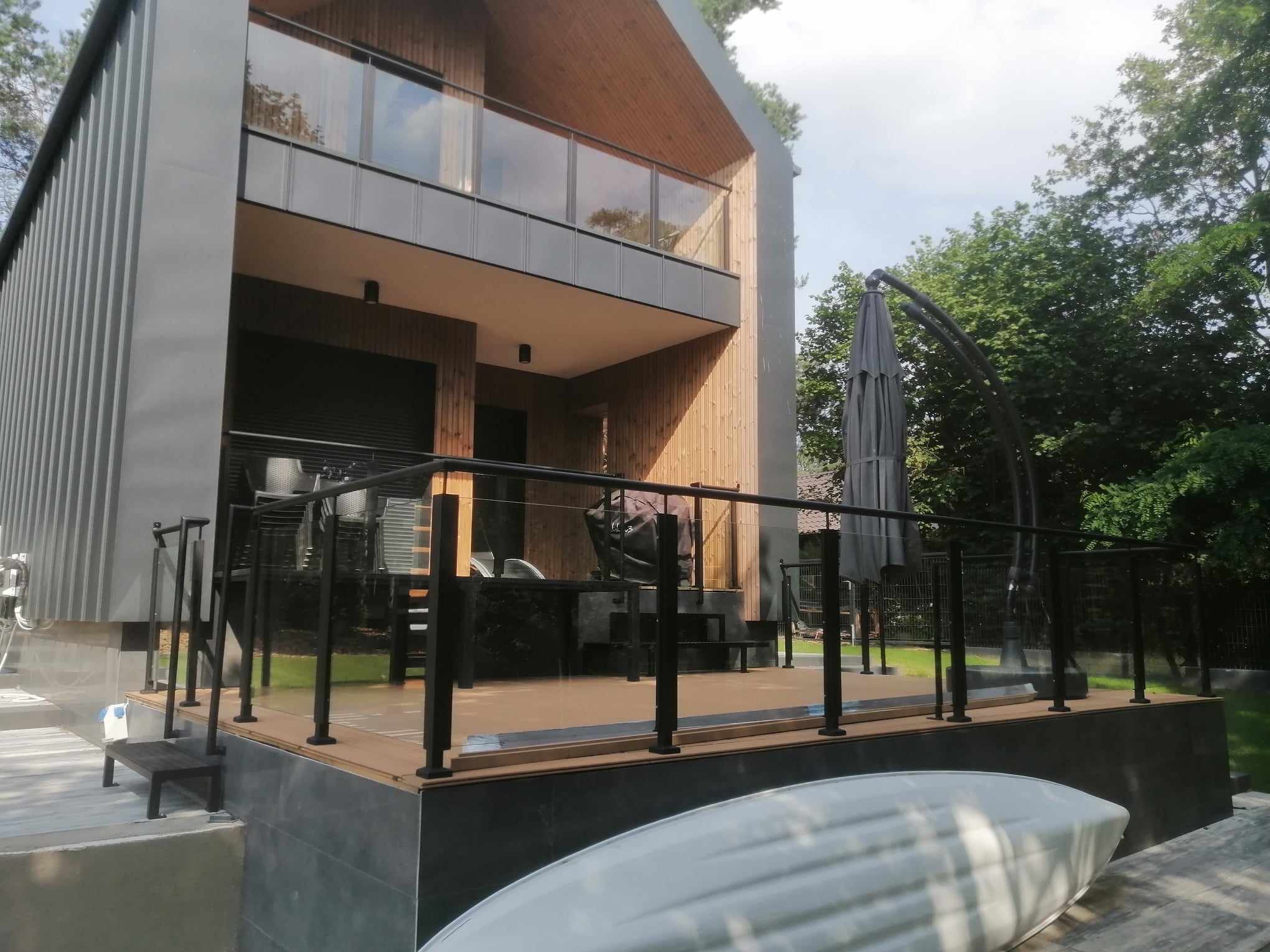 Nowoczesny dom z drewnianą elewacją i balkonem zabezpieczonym szklaną balustradą z czarnymi słupkami, widok na taras z czarną balustradą i meblami ogrodowymi, częściowo zasłonięty przez rozłożony...