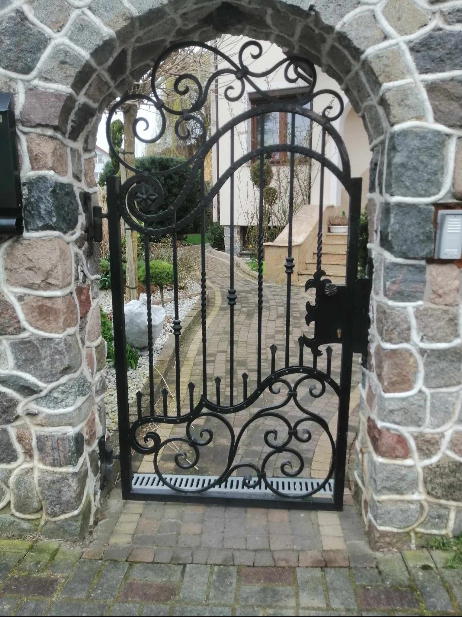 Otwarta, ozdobna, czarna brama z kutego żelaza, osadzona w kamiennym łuku wjazdowym, prowadząca do ogrodu i domu.