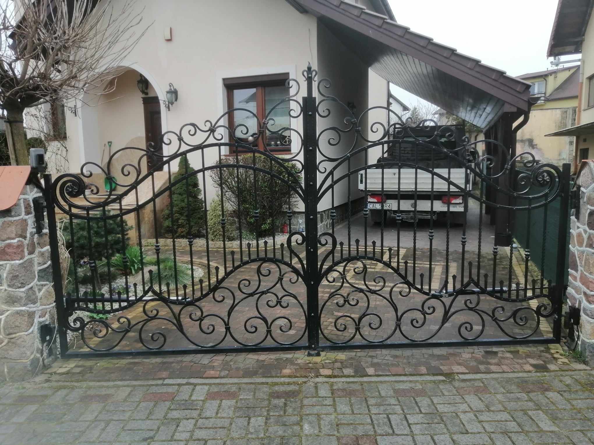 Ozdobna, dwuskrzydłowa brama z kutego żelaza, z motywami roślinnymi, prowadząca do posesji z beżowym domem i wiatą garażową, widziana z poziomu brukowanego podjazdu.