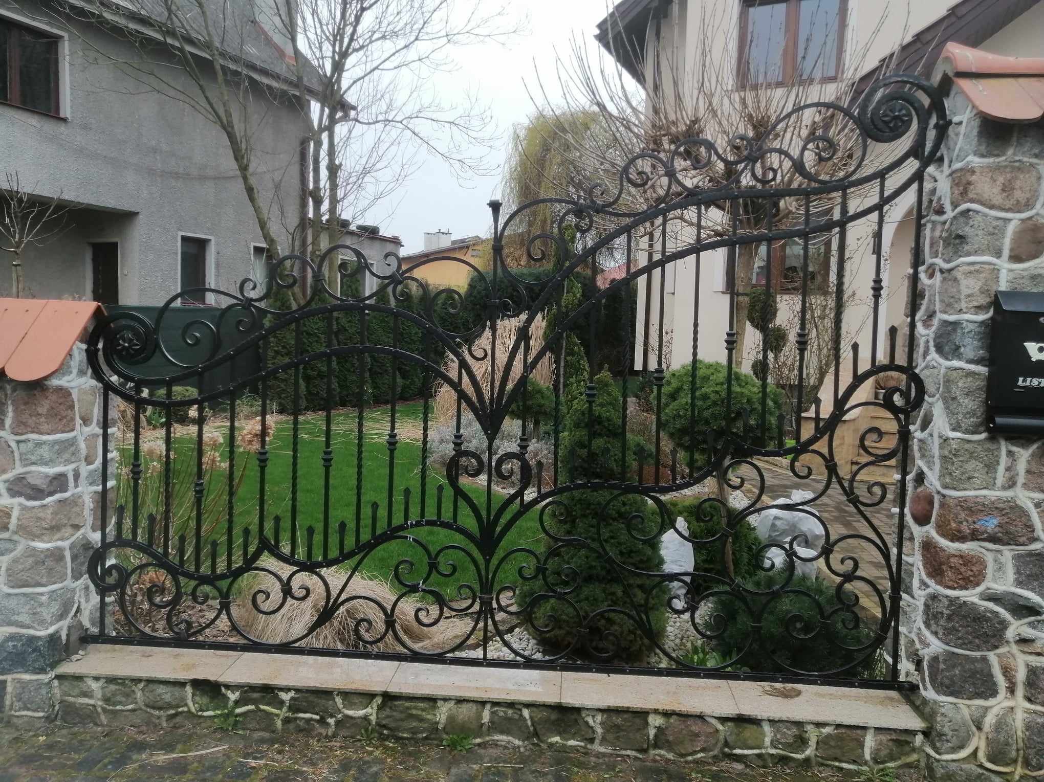 Ozdobna, czarna brama kuta z motywami roślinnymi, zamontowana na kamiennych słupkach, z widokiem na zadbany ogród i domy w tle.