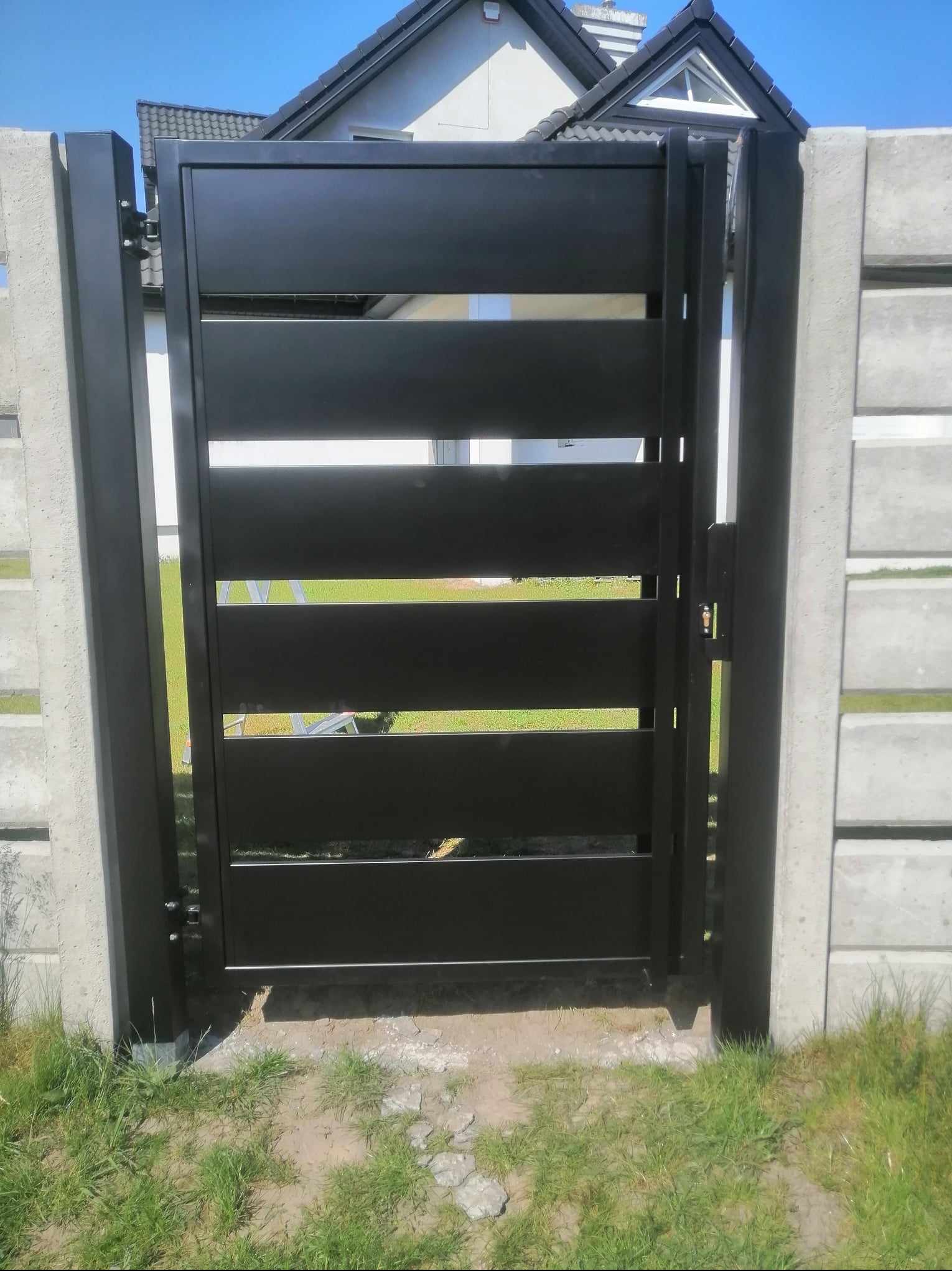 Nowoczesna, czarna brama wjazdowa z poziomymi panelami, zamontowana pomiędzy betonowymi słupkami ogrodzeniowymi, widoczna z bliska w słoneczny dzień.