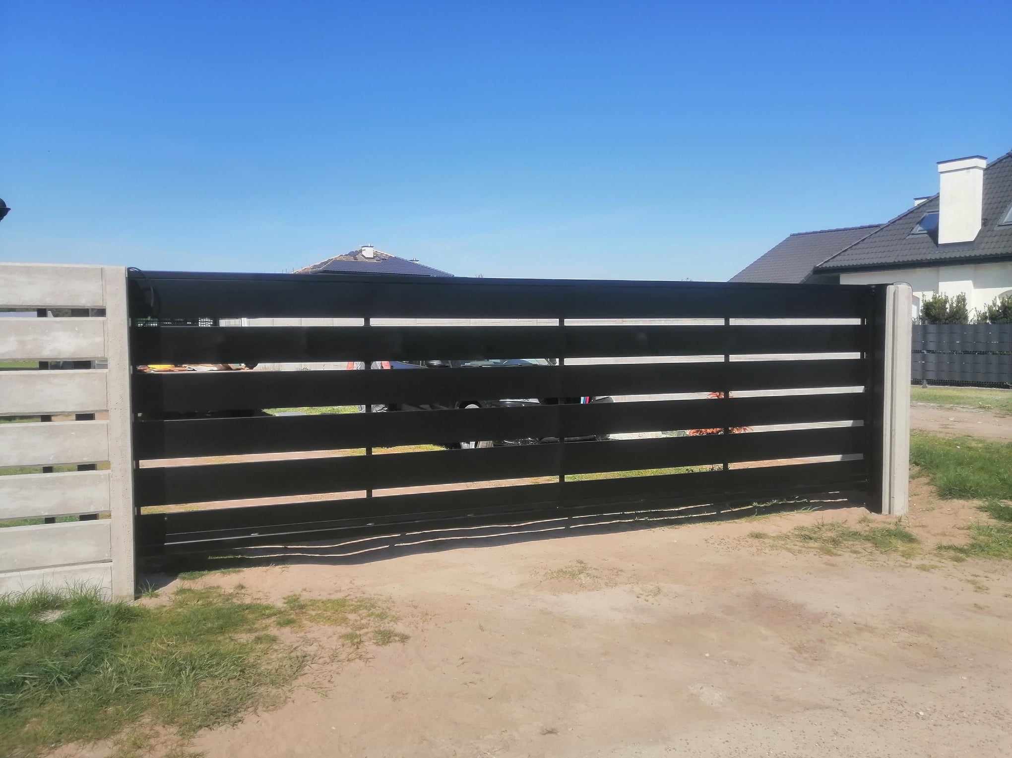 Nowoczesna, przesuwna brama z poziomych, czarnych profili, osadzona na betonowych słupkach, na tle błękitnego nieba i zabudowy jednorodzinnej.
