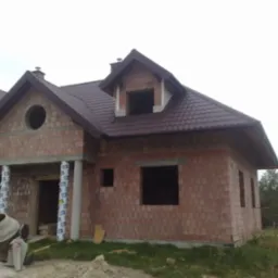 Surowy, dwukondygnacyjny dom z czerwonej cegły w trakcie budowy, z widoczną mieszarką do betonu i oknami bez ram.