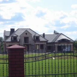 Niedokończony, dwukondygnacyjny dom z cegły z ciemną dachówką, widoczny przez czarne, metalowe ogrodzenie z ozdobnymi elementami, na tle zielonego trawnika i błękitnego nieba z chmurami.