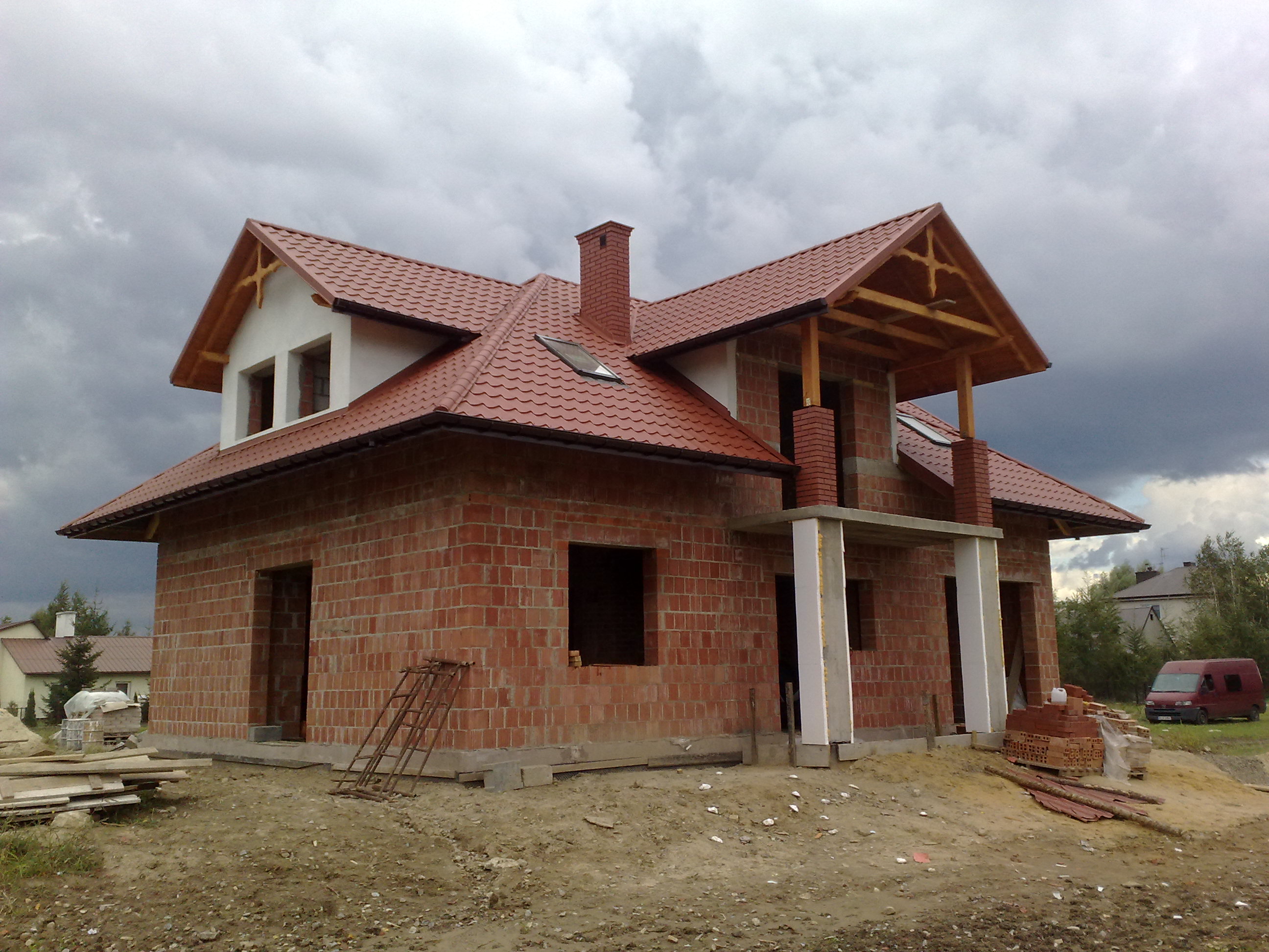 Surowy, dwukondygnacyjny dom z czerwonej cegły w trakcie budowy, z częściowo zamontowaną czerwoną dachówką, drewnianą konstrukcją werandy i ponurym, zachmurzonym niebem w tle.