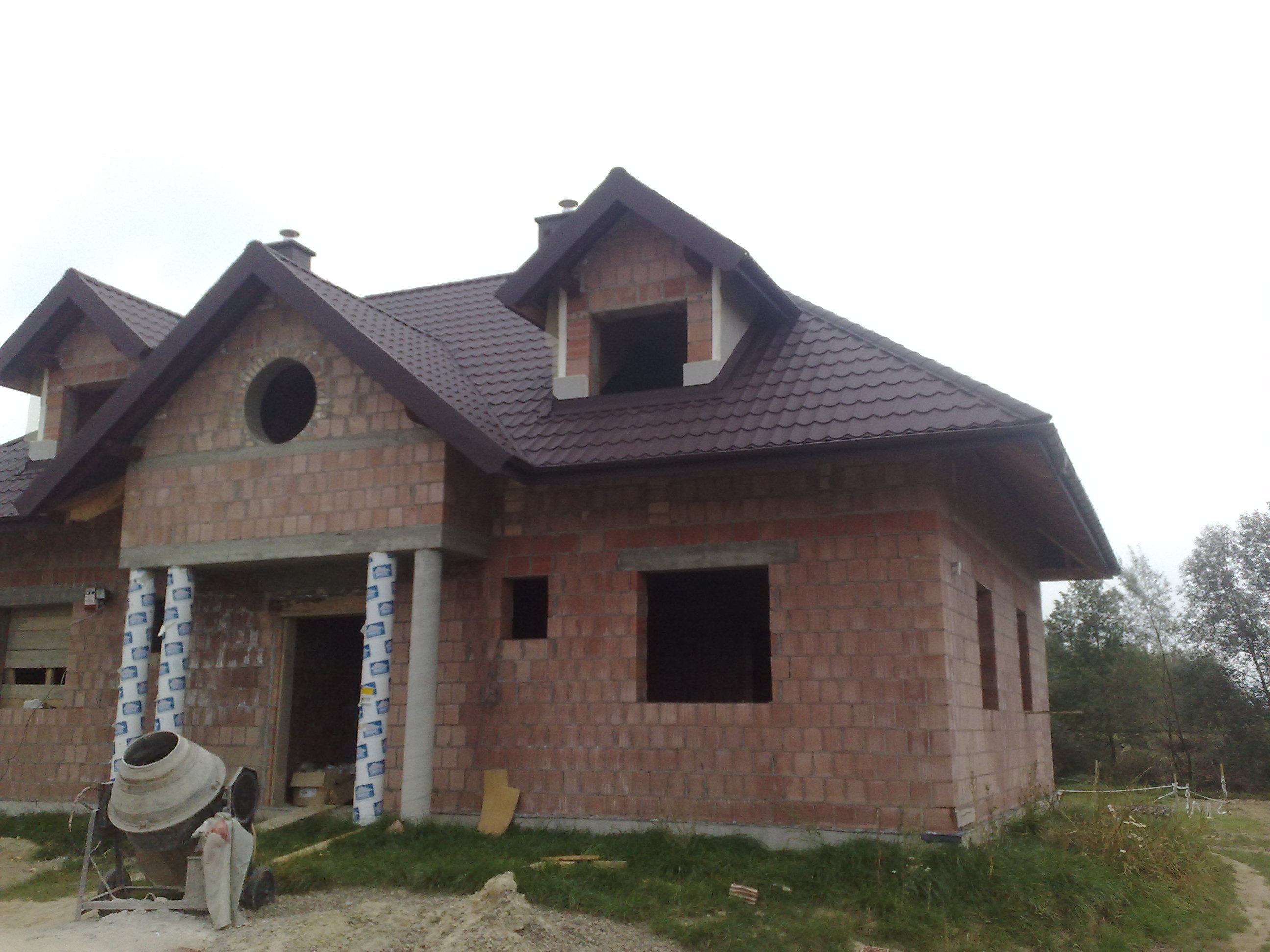 Surowy, dwukondygnacyjny dom z czerwonej cegły w trakcie budowy, z widoczną mieszarką do betonu i oknami bez ram.