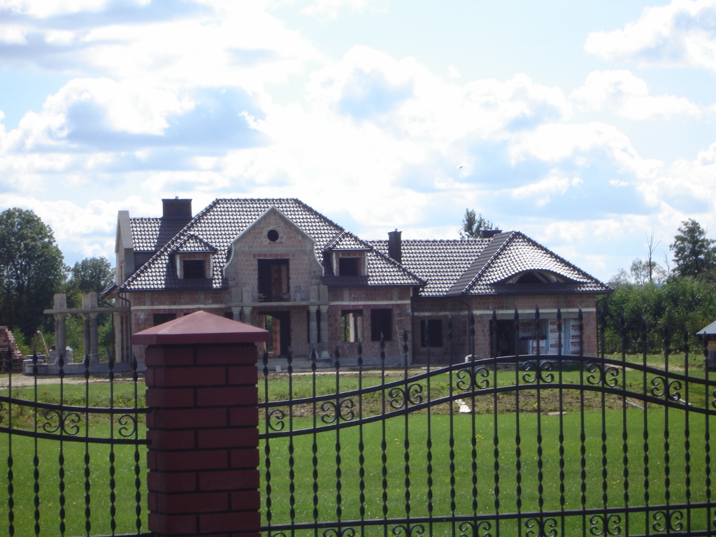 Niedokończony, dwukondygnacyjny dom z cegły z ciemną dachówką, widoczny przez czarne, metalowe ogrodzenie z ozdobnymi elementami, na tle zielonego trawnika i błękitnego nieba z chmurami.
