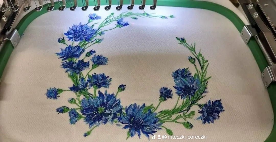 Haftowany wieniec z chabrów na białym materiale w tamborku maszyny hafciarskiej.