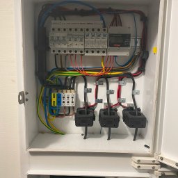 ADC INSTALACJE ELEKTRYCZNE GRZEGORZ ADAMCZYK - Otwarta skrzynka rozdzielcza z zainstalowanymi bezpiecznikami, licznikiem energii Fronius Smart Meter i przewodami w różnych kolorach, widoczne również trzy przekładniki prądowe.