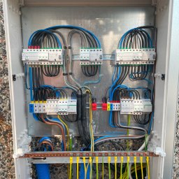 ADC INSTALACJE ELEKTRYCZNE GRZEGORZ ADAMCZYK - Otwarta skrzynka rozdzielcza z zainstalowanymi bezpiecznikami, przewodami w różnych kolorach i niebieskimi rurkami karbowanymi, zamontowana na zewnątrz budynku z elewacją z drobnych kamieni.