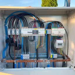 ADC INSTALACJE ELEKTRYCZNE GRZEGORZ ADAMCZYK - Otwarta skrzynka elektryczna z widocznymi przewodami, zabezpieczeniami i licznikiem energii, zamontowana na zewnątrz budynku, z zielonymi drzewami w tle.