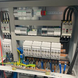 ADC INSTALACJE ELEKTRYCZNE GRZEGORZ ADAMCZYK - Otwarta skrzynka rozdzielcza z zainstalowanymi bezpiecznikami, wyłącznikami i przewodami o różnych kolorach, widoczne elementy montażowe i oznaczenia na urządzeniach.