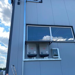 ADC INSTALACJE ELEKTRYCZNE GRZEGORZ ADAMCZYK - Dwa inwertery marki Fronius ECO zamontowane na elewacji budynku, widoczne okablowanie, drabina oparta o ścianę, błękitne niebo z chmurami odbija się w szybie okna.