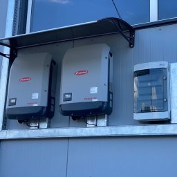 ADC INSTALACJE ELEKTRYCZNE GRZEGORZ ADAMCZYK - Dwa inwertery Fronius ECO zamontowane na zewnątrz budynku z szarej blachy trapezowej, pod czarnym zadaszeniem, obok skrzynka elektryczna z transparentną pokrywą.