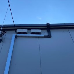 ADC INSTALACJE ELEKTRYCZNE GRZEGORZ ADAMCZYK - Dwa falowniki fotowoltaiczne zamontowane na elewacji budynku, widoczne czarne rury kablowe, kamera przemysłowa i maszt odgromowy na tle błękitnego nieba.