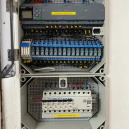 Otwarta szara szafka elektryczna z widocznymi elementami sterowania oświetleniem, w tym sterowniki Siemens LOGO!, przekaźniki i zabezpieczenia Eaton, zamontowana na białej ścianie o wyraźnej fakturze.