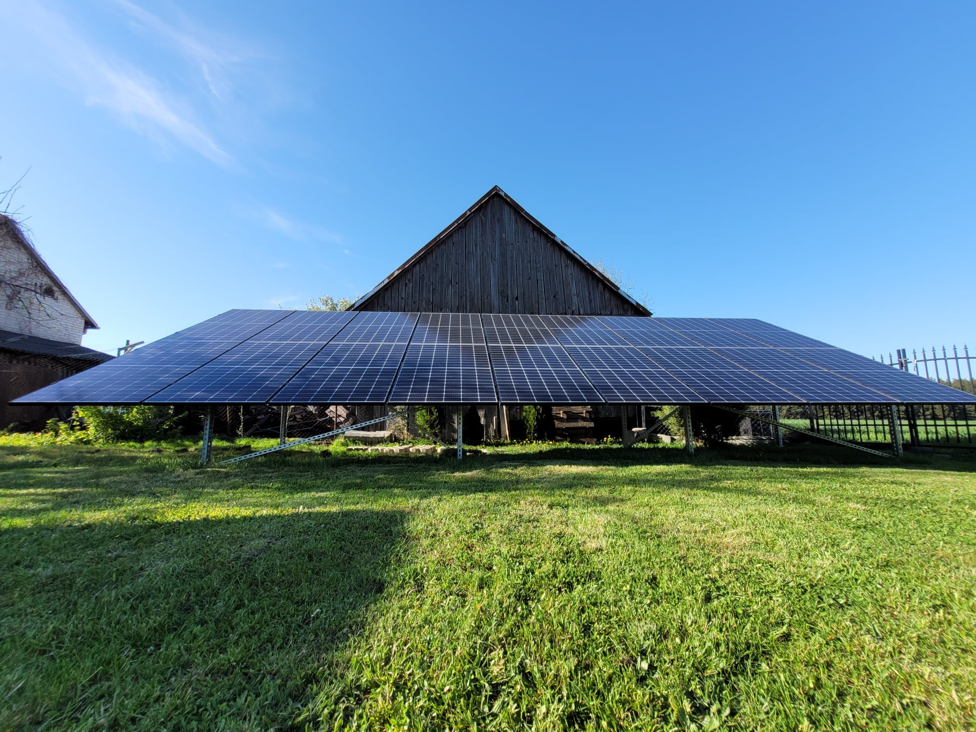 Instalacja paneli słonecznych na konstrukcji wsporczej na trawniku, z drewnianym budynkiem w tle pod błękitnym niebem.