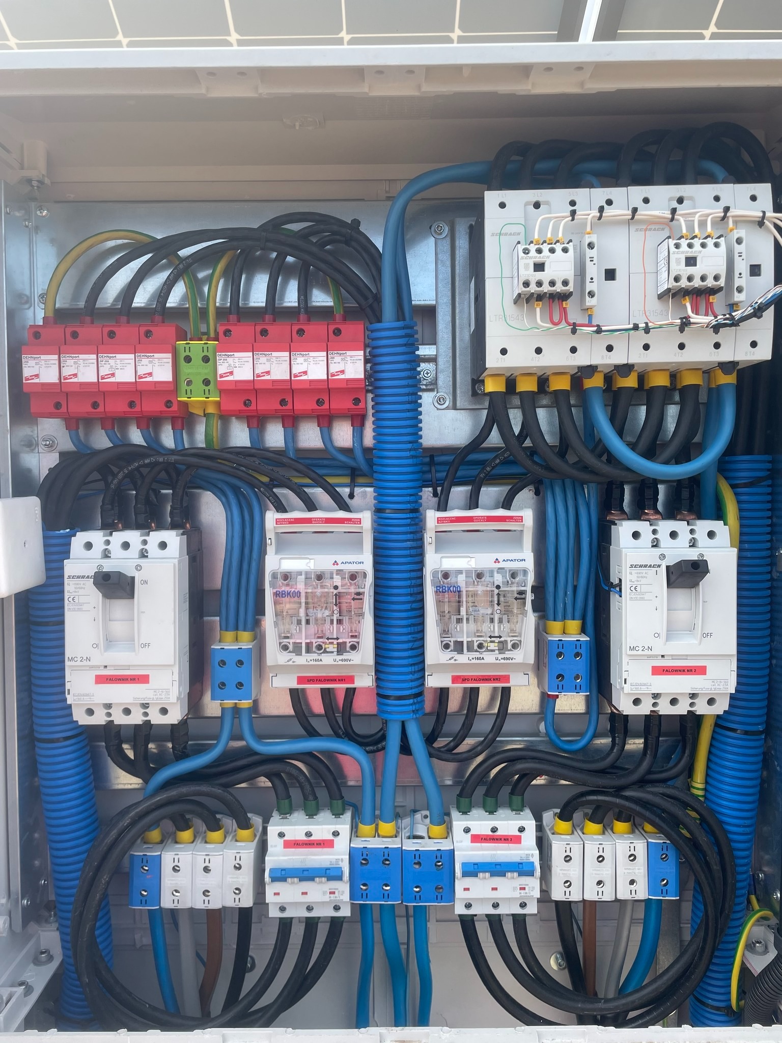 Rozdzielnica elektryczna z elementami zabezpieczeń przepięciowych, bezpiecznikami i przewodami w niebieskich peszlach, instalacja fotowoltaiczna.