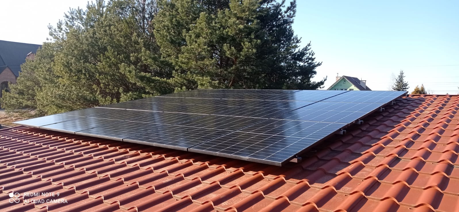 Panele słoneczne zamontowane na czerwonym dachu z dachówki ceramicznej, z widocznymi drzewami iglastymi i fragmentami budynków w tle, jasne niebo.