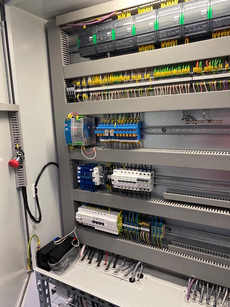 Otwarta szafa sterownicza z zainstalowanymi modułami PLC, bezpiecznikami, przekaźnikami i listwami zaciskowymi, okablowanie uporządkowane, widoczne przewody w kolorowych izolacjach.