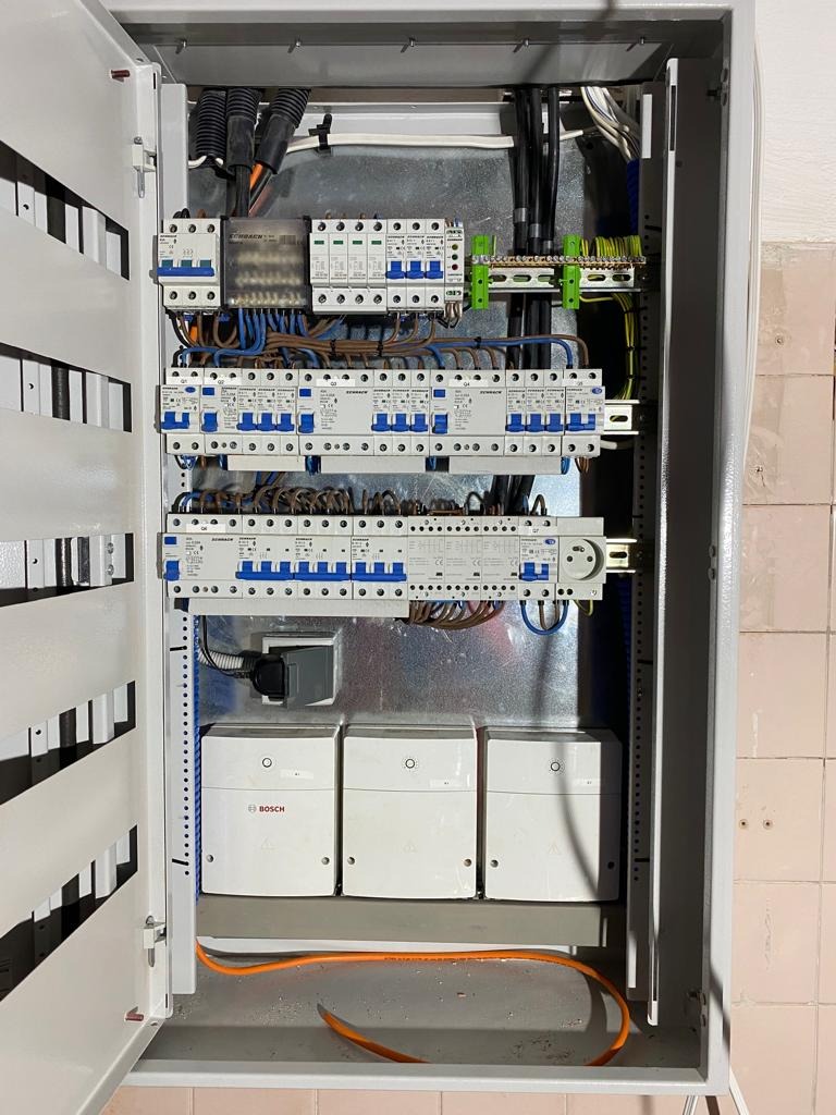 Otwarta skrzynka elektryczna z wieloma bezpiecznikami, przewodami i trzema białymi modułami Bosch na dole, widoczna na tle ściany z ceramicznych płytek.