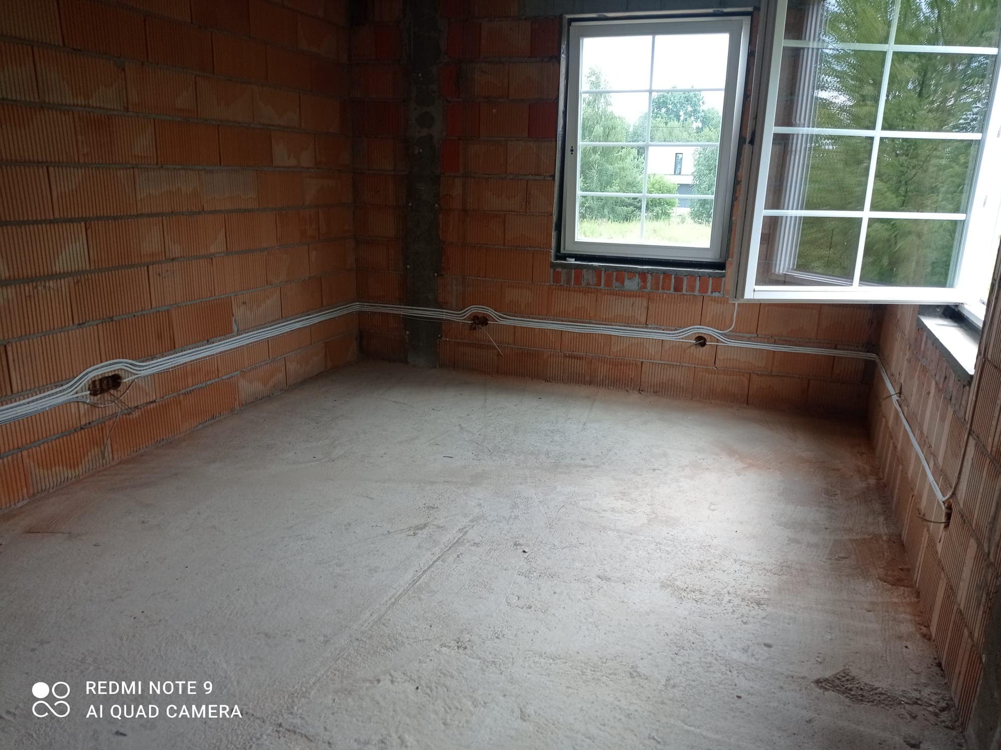 Surowy stan wnętrza z ceglanymi ścianami, widoczne poprowadzone przewody elektryczne w białych listwach i otwarte okno z widokiem na zieleń.