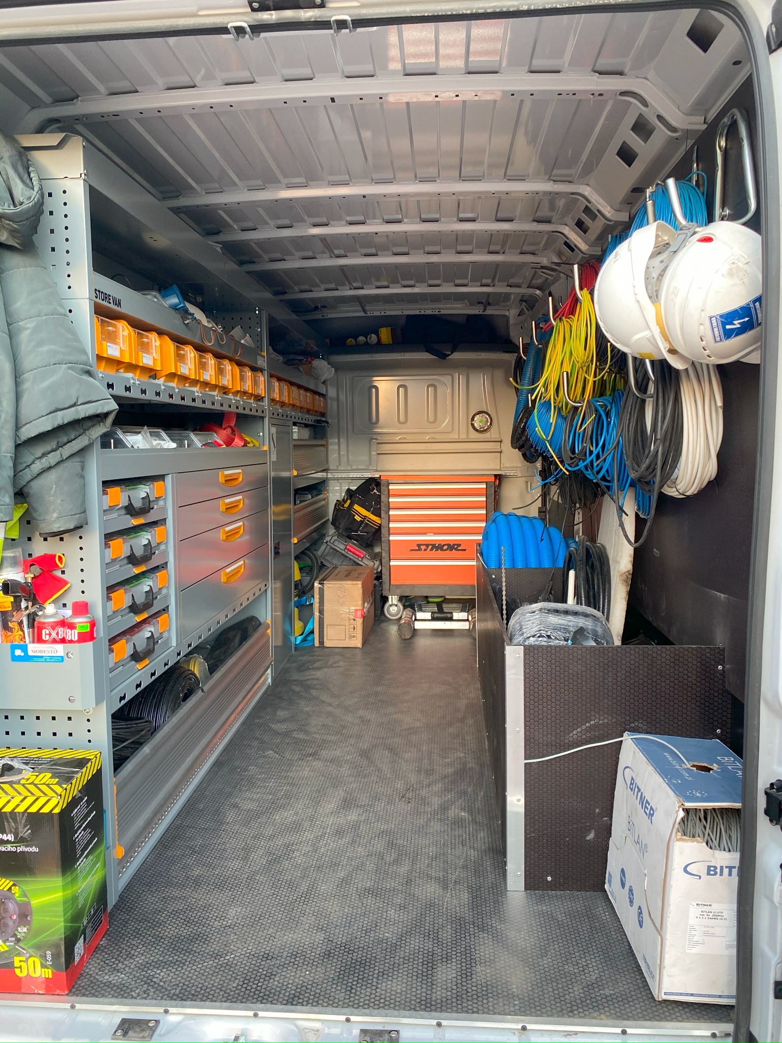 Wnętrze furgonetki serwisowej z regałami i szufladami wypełnionymi narzędziami i materiałami elektrycznymi, w tym kable, puszki, skrzynka narzędziowa Sthor i kaski ochronne z symbolami elektrycznymi.