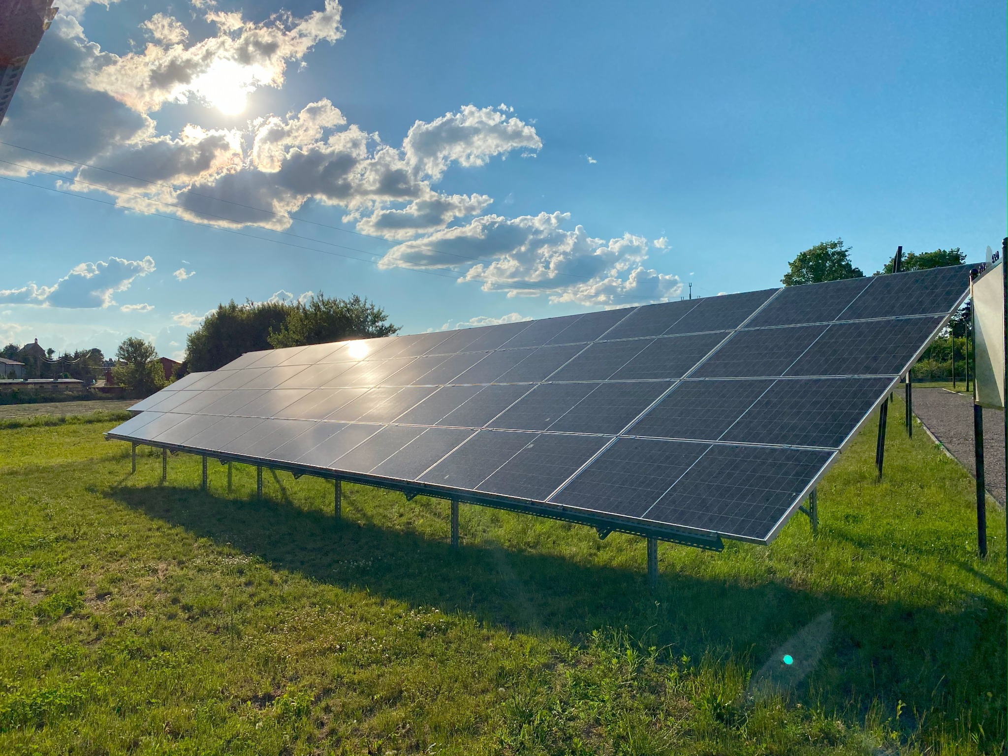 Rząd paneli słonecznych na metalowej konstrukcji, odbijających światło słoneczne, na tle zielonej trawy, błękitnego nieba z chmurami i drzew w oddali.