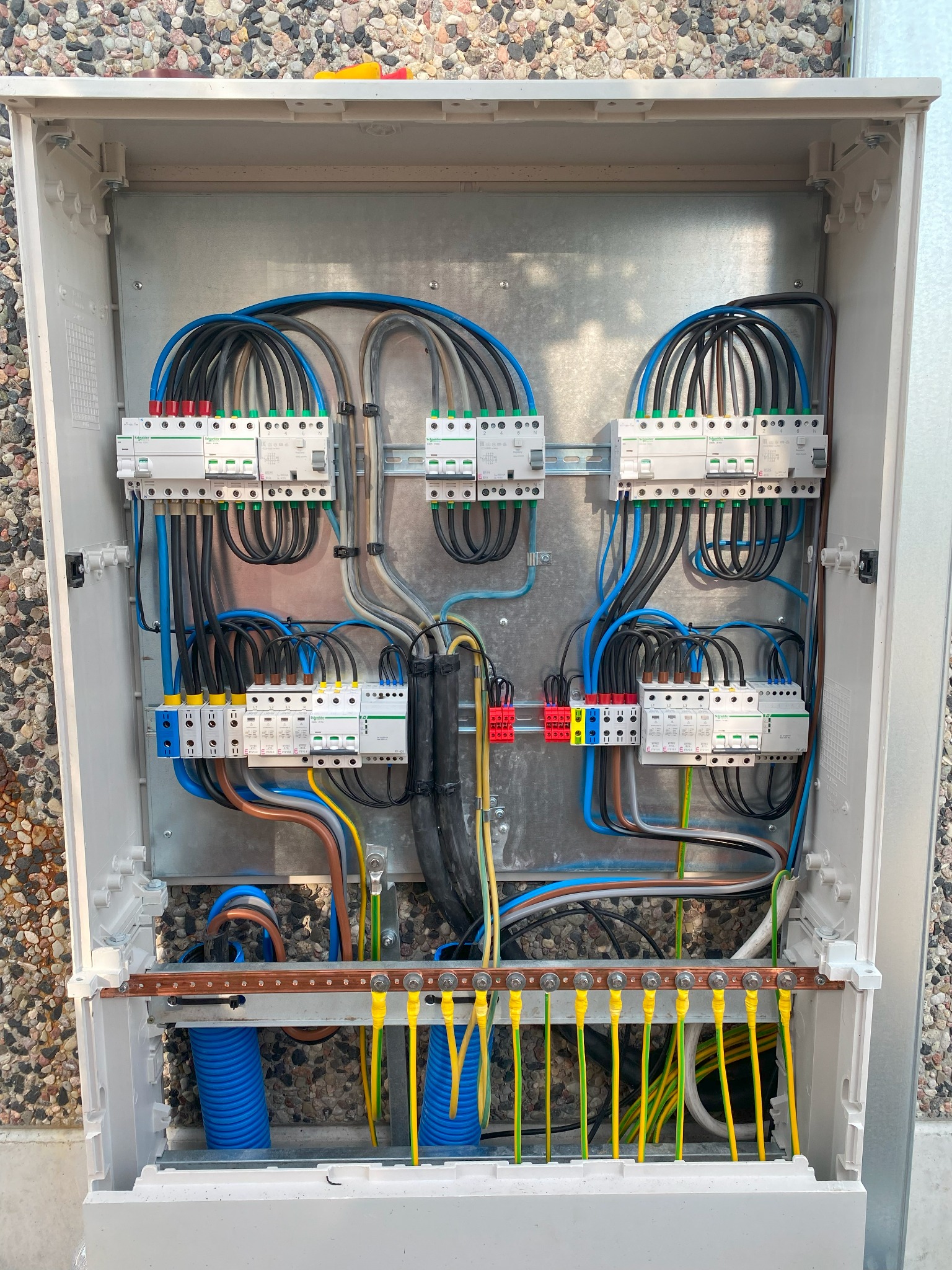 Otwarta skrzynka rozdzielcza z zainstalowanymi bezpiecznikami, przewodami w różnych kolorach i niebieskimi rurkami karbowanymi, zamontowana na zewnątrz budynku z elewacją z drobnych kamieni.
