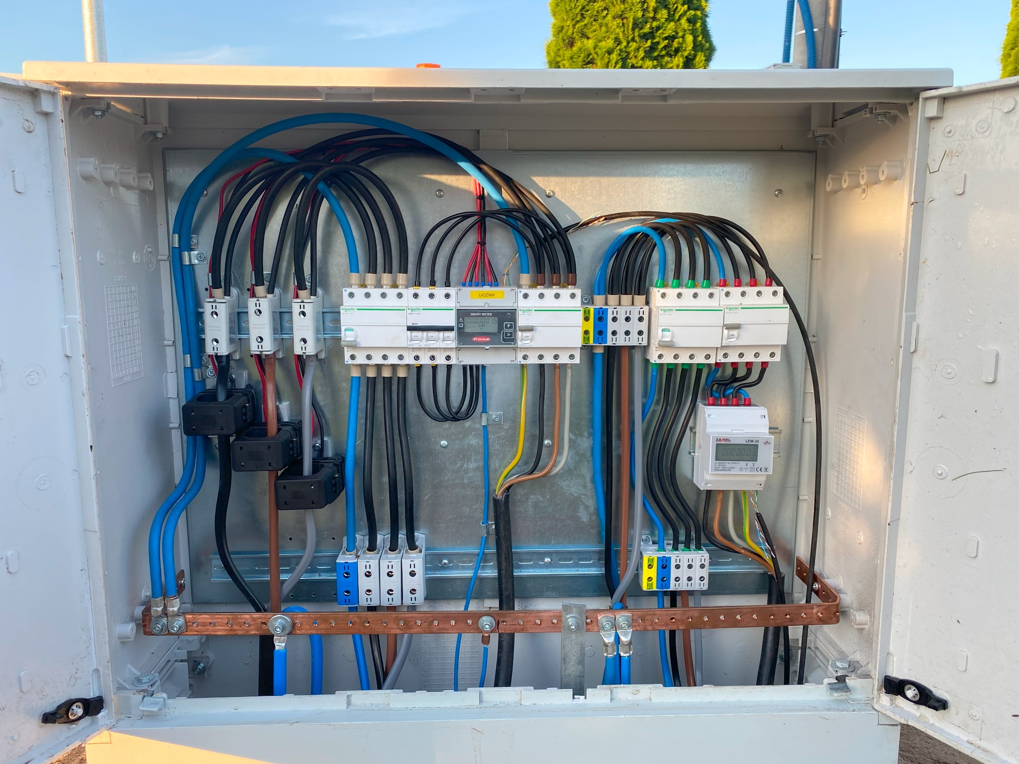 Otwarta skrzynka elektryczna z widocznymi przewodami, zabezpieczeniami i licznikiem energii, zamontowana na zewnątrz budynku, z zielonymi drzewami w tle.