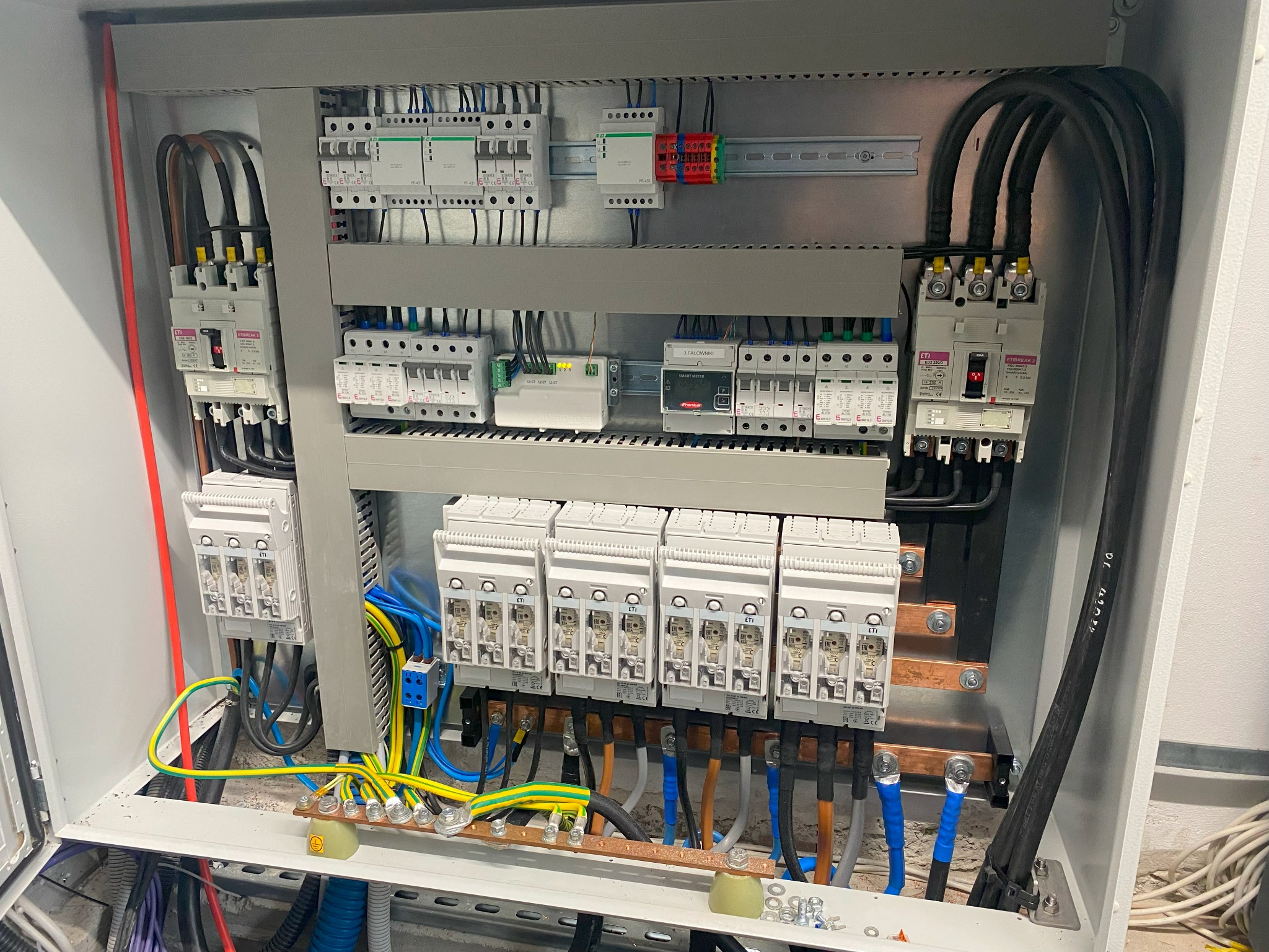 Otwarta skrzynka rozdzielcza z zainstalowanymi bezpiecznikami, wyłącznikami i przewodami o różnych kolorach, widoczne elementy montażowe i oznaczenia na urządzeniach.