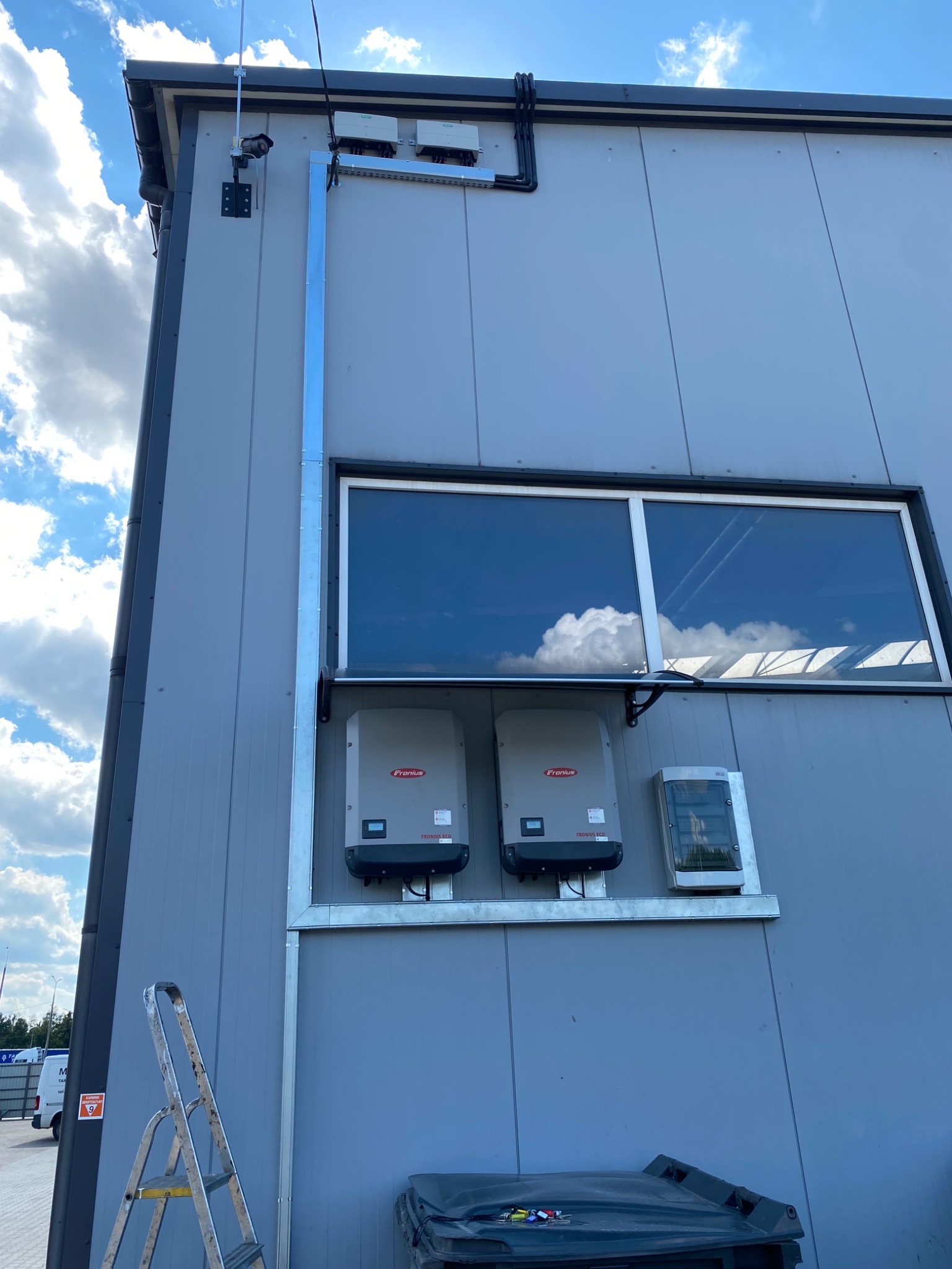 Dwa inwertery marki Fronius ECO zamontowane na elewacji budynku, widoczne okablowanie, drabina oparta o ścianę, błękitne niebo z chmurami odbija się w szybie okna.