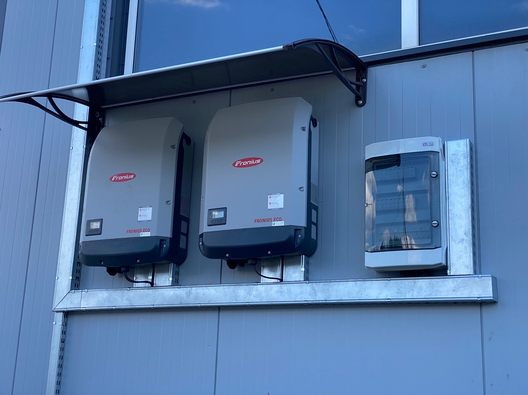 Dwa inwertery Fronius ECO zamontowane na zewnątrz budynku z szarej blachy trapezowej, pod czarnym zadaszeniem, obok skrzynka elektryczna z transparentną pokrywą.